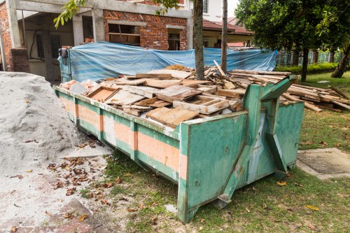 A large metal skip with a turquoise finish, positioned outdoors on a grassy area near a residential street in Pinner, containing a substantial pile of weathered wooden planks and panels with rough, uneven surfaces and a mix of brown and grey tones. The skip's sides have orange reflective strips, and it is partially filled with construction or renovation debris, with some wooden pieces leaning against the sides. In the background, there is a brick building under construction or renovation, covered with blue tarpaulin, and nearby trees and grass contribute to the suburban environment. The scene suggests clearance or removal of building materials, aligning with rubbish removal services provided by Office Clearance Pinner in the local area.