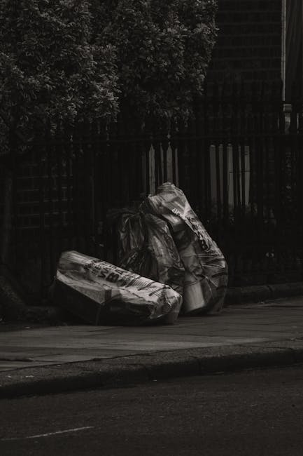 An outdoor scene in front of a commercial building showing overflowing waste bins and numerous discarded rubbish bags, boxes, and packaging materials scattered on the paved ground and partially spilli