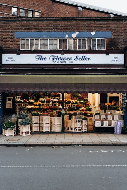 The image depicts the storefront of a flower shop named 'The Flower Seller of Muswell Hill,' located on a street with a brick building above. The shop's sign, painted in a light script font, displays 