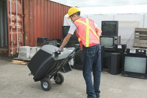 A waste disposal professional wearing a yellow safety helmet, high-visibility vest with reflective strips, and casual navy trousers is seen in an outdoor area near a red shipping container and a makeshift shelter. The worker is pushing a black wheeled trolley loaded with a large, dark-colored plastic rubbish bin. Behind them, there is a collection of old electronic appliances, including at least three CRT televisions and several black electronic devices, placed against the container and on wooden pallets. The surface appears to be a paved yard or lot with a mix of metal and wood materials, suitable for waste collection and disposal activities for businesses or offices in Pinner. The scene is well-lit, with natural daylight illuminating the objects clearly, aligning with professional rubbish removal services offered by companies like Office Clearance Pinner.