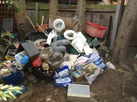 A confined outdoor space with a cluttered pile of miscellaneous rubbish, including white plastic pipes, a black plastic container, crumpled paper and plastic bags, cardboard boxes, and various discarded household items, all situated on the ground near a wooden fence and several tree trunks. The debris appears to be a mixture of construction waste, packaging, and general household refuse, with some items leaning against or partially covered by others. The scene is illuminated by natural daylight, highlighting the disorganized collection of rubbish which is typical of a site requiring waste clearance services in Pinner or nearby areas, as managed by Office Clearance Pinner. The surrounding environment includes a patch of bare earth and the base of trees, with the fence and wooden posts providing a backdrop.